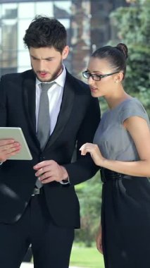 Kendine güvenen bir işadamı ve başarılı bir iş kadını, işbirliklerinde bir dönüm noktası teşkil eden heyecan verici haberlere dalmış, açık havada bir tabletle neşeyle meşgul oluyorlar. Şık, modern kıyafetleri yemyeşil ve yemyeşil ile çok güzel zıtlaşıyor.