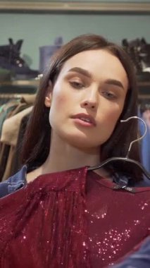 Göz kamaştırıcı genç bir kadın, şık bir butik soyunma odasında göz kamaştırıcı bir elbise tutarak kameraya güvenle gülümsüyor. Bir dizi şık kıyafetle çevrelenmiş, mükemmel kıyafeti bulmanın heyecanını yaşıyor, neşe ve zarafet saçıyor.