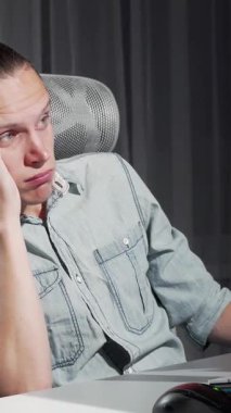 Sıradan açık mavi bir gömlek giymiş yorgun bir genç adam, ergonomik sandalyesinde yığılmış masasında uykuya dalıyor. Bilgisayar ekranının yumuşak parıltısı loş ışıklı odayla çelişiyor.