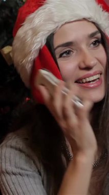 Neşeli, beyaz bir kadın Noel Baba şapkası takmış cep telefonuyla sohbet ederken etrafı renkli süsler ve parlak ışıklarla süslenmiş çarpıcı bir Noel ağacıyla çevrili. Sıcak gülüşü ve rahat süveteri bayram neşesi saçıyor.