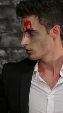 Gösterişli siyah takım elbiseli büyüleyici bir erkek vampir koyu renkli tuğla bir duvara karşı dramatik bir poz veriyor. Alnında kan kırmızısı bir aksan olan çarpıcı makyajı, Cadılar Bayramı 'nın ürkütücü cazibesini arttırıyor.