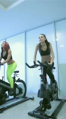 Hareketli bir sahne, iki kararlı arkadaşın hareketsiz bisikletlerle pedal çevirmesini, ifadelerinin odak noktasını ve yoğunluğunu parlak bir spor salonunda yansıtmasını çeker. Moda spor kıyafetleri giyip, enerji ve motivasyon yayıyorlar.