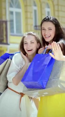 Üç coşkulu kız, her biri şık kıyafetler giymiş, hareketli bir alışveriş bölgesinin dışında canlı alışveriş torbalarını tutarken coşkulu bir şekilde el sallıyorlar. Işıl ışıl gülüşleri ve tasasız enerjileri arkadaşlığın neşesini ve heyecanı özetliyor.