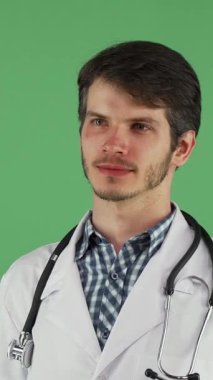 Çıtır beyaz önlüklü ve kareli gömlekli genç bir doktor sıcak bir gülümseme sergiliyor, kendine güven ve yakınlık yayıyor. Boynunda zarif bir steteskop ile canlı yeşil bir kromakey arka planına karşı duruyor.