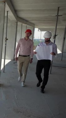 İki erkek müteahhit, biri parlak beyaz kasklı, diğeri parlak turuncu kasklı, yüksek katlı bir binanın kenarında, özenle planlarını inceliyor. Nefes kesen şehir silüeti ve yemyeşil altta dinamik bir hava yaratır.