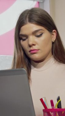 Kararlı bir kız öğrenci, gözle görülür şekilde yorgun ve stresli, renkli masasında oturur, dizüstü bilgisayarına dikkatle odaklanırken nazikçe şakaklarını ovuşturur. Sıcak, yumuşak ışık, onun güzelliğini ve akademik baskının gerilimini vurguluyor.