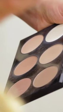 Yakından çekilen bir fotoğraf bir kadının ellerini ustalıkla yakalar parlak bir paletten canlı bir ruj sürerek özgüven ve sanatsallık yayar. Kuaför salonunun enerjik atmosferi dönüşümün heyecanını arttırır ve güzelliğin karmaşıklığını ön plana çıkarır.