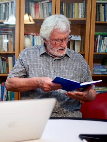 Portrait of a white bearded senior man with grey shirt reading a blue notebook, book while sitting in front of a huge book shelf in the background.