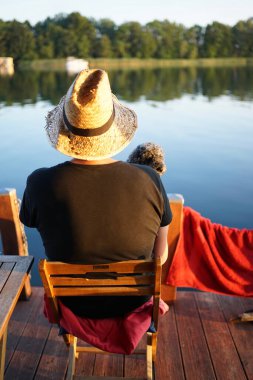 Hasır şapkalı ve köpekli bir adam gün batımında sakin gölü seyrediyor.            