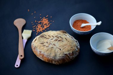 Kara tahtada ev yapımı buğday ekmeği, arka planda bir fincan kırmızı leniles ile koyu kabuklu ekşi ekmek.