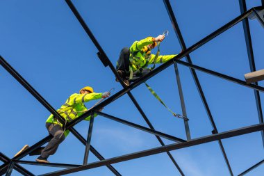 İnşaat alanında güvenlik yüksekliği ekipmanı; Asyalı işçiler çatıyı kurmak için güvenlik yüksekliği ekipmanı takıyorlar. Emniyet kemeri için kancaları olan işçiler için tutuklama aygıtını düşür.