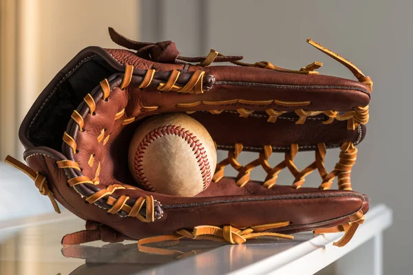 Close-up of a used baseball ball inside brown, leather baseball glove on plain, grey background.
