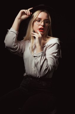 Stüdyoda dramatik ışık ile poz çıplak omuz ile gömlek giyen lüks genç kadın gözlük ve şapka,