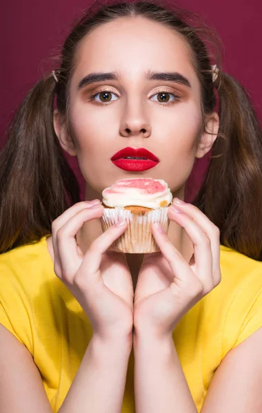 Pretty brunette model with bright makeup holding sweet cupcake. Studio shot on a red background