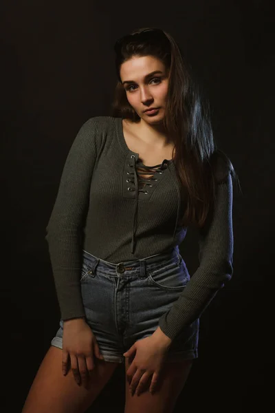 Fashion model test shoot of attractive woman posing in dark studio dressed in grey underwear and jeans shorts