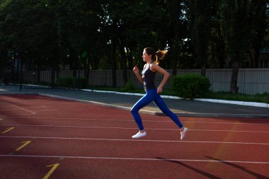 Gri t gömlek ve mavi tozluk güneşli gece boyunca stadyum yolda çalışan genç atletik kız. Metin için yer