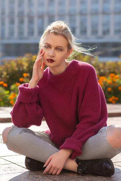 Pretty blonde woman wearing red oversize sweater sitting on the paving tiles