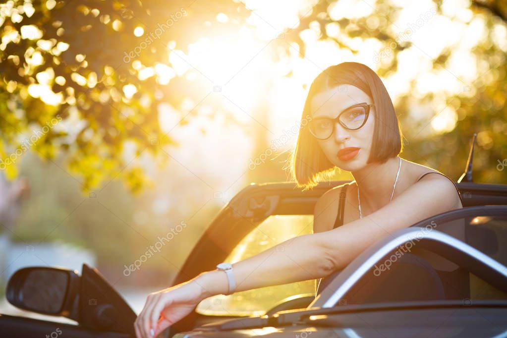 Stylish looking brunette woman with red lips wearing glasses, posing in cabriolet with a sun light. Empty space