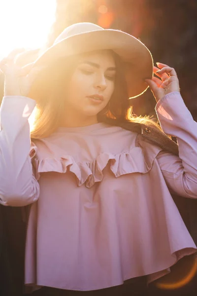 Elegant tanned woman wearing hat and blouse posing in rays of sun