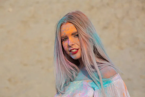 Closeup portrait of stunning blonde woman playing with blue dry Holi paint at the desert