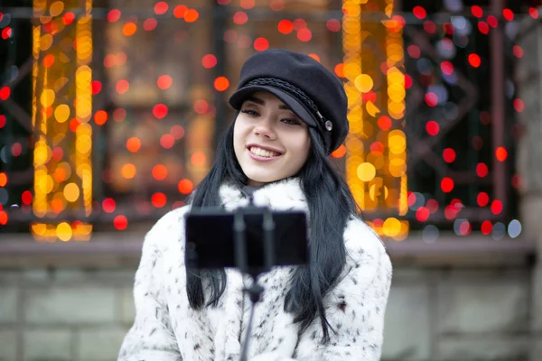 Lovely brunette woman wearing cap, taking self portrait at the street with blurred lights