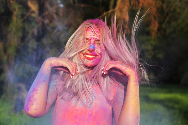 Adorable blonde woman with fluttering hair posing covered of a colorful paint at the Holi Festival