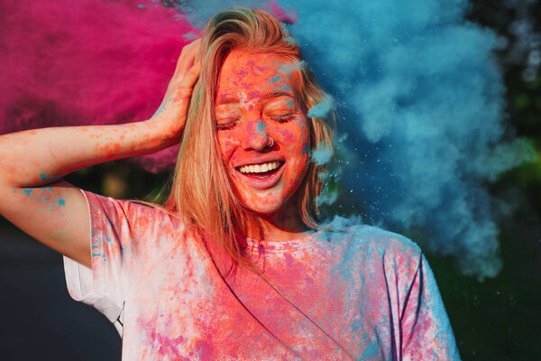 Позитивная блондинка веселится со взрывающимся голубым и розовым сухим порошком, празднующим фестиваль Холи
