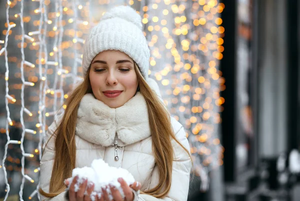 光彩照人的金发碧眼的女人戴着帽子和温暖的外套 手里拿着雪 在花环模糊的背景 空空间 — 图库照片