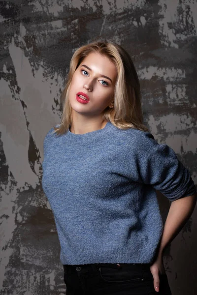 Stunning blonde woman with lush hair wearing blue sweater, posing in studio with shadows