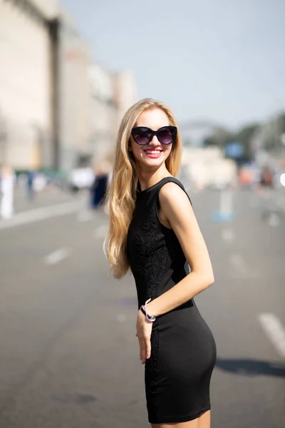 神话般的微笑的女人穿着黑色礼服和太阳镜 走在阳光明媚的街道上 — 图库照片