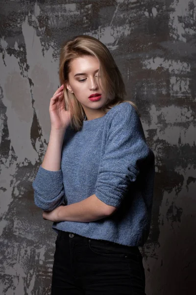 Lovely blonde woman with lush hair wearing blue sweater, posing in studio with shadows