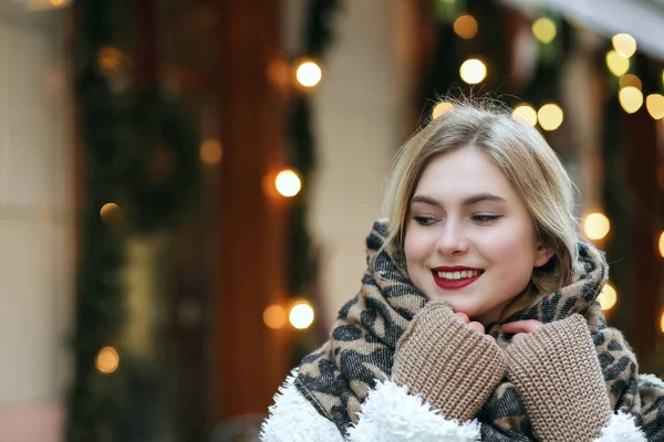 Joyful blonde woman wears winter coat and knit scarf walking at the city. Empty space