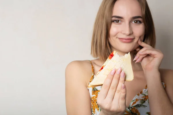 Χαμογελαστή μελαχρινή κορίτσι φοιτητής τρώει ένα σάντουιτς πάνω από μια γκρίζα πλάτη — Φωτογραφία Αρχείου