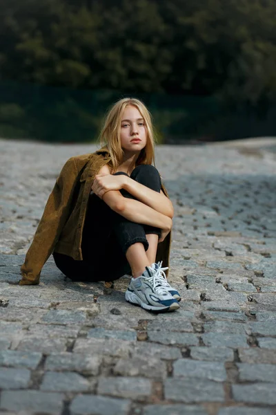 Triste menina loira sentada na estrada com pedras de pavimentação — Fotografia de Stock