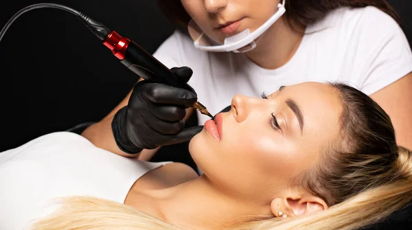 Woman in gloves and plastic mask making permanent lip makeup to a pretty blonde woman in cosmetology salo