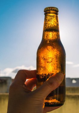 Arka planda gökyüzü olan soğuk bir bira şişesini tutarken