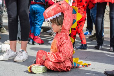 Dinozor kostümlü çocuk etrafı insanlarla çevrili yerde oturuyor. Küçük bir çocuk Cadılar Bayramı için hazırlanıyor..