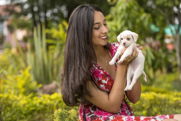 Beautiful young latin woman in the park with a small white puppy in her hands.