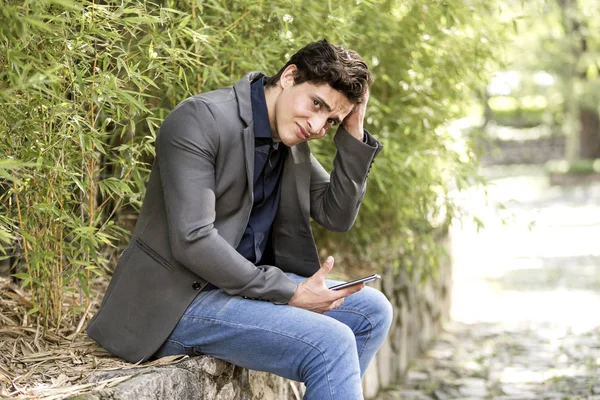 Young troubled businessman sitting in garden with cell phone in his hand.