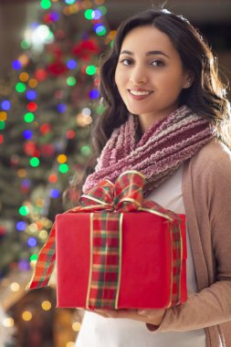 Kırmızı hediye ve Noel ağacı içinde belgili tanımlık geçmiş ile gülümseyen genç güzel latin kadın portresi