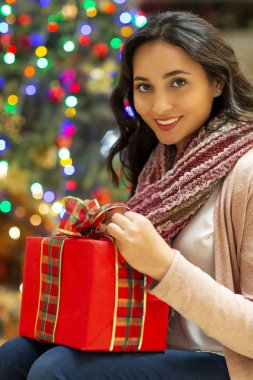 Kırmızı hediye ve Noel ağacı içinde belgili tanımlık geçmiş ile gülümseyen genç güzel latin kadın portresi