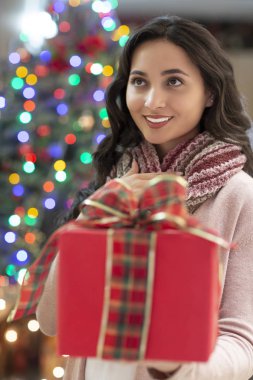 Kırmızı hediye ve Noel ağacı içinde belgili tanımlık geçmiş ile gülümseyen genç güzel latin kadın portresi