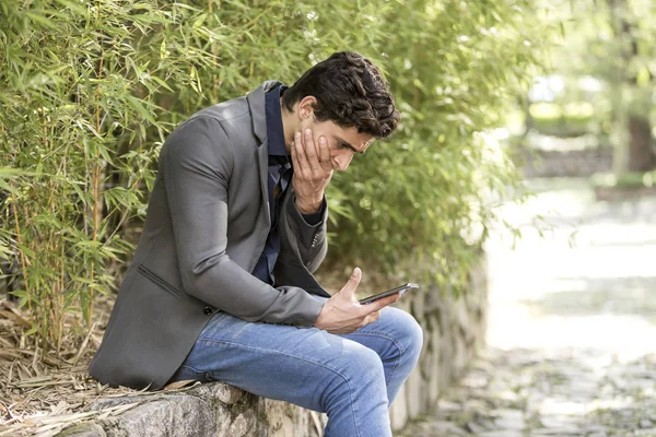 Young troubled businessman sitting in garden with cell phone in his hand.