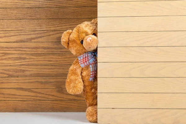 Teddy bear standing indoors with a wooden floor and wall.