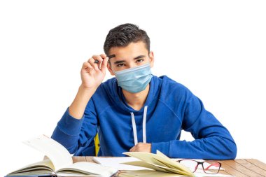 Maskeli genç latin adam dizüstü bilgisayarlı bir masada çalışıyor ve beyaz arka plandaki kameraya bakıyor.. 