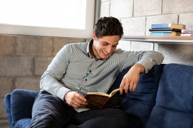 Yakışıklı ve zarif bir adam oturma odasında COVID-19 salgını sırasında kitap okuyor.