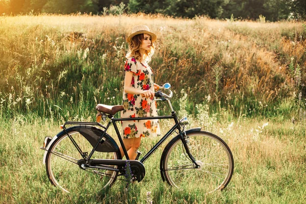 Romantic hipster girl on retro bike in the field, dressed in light summer dress with flowers and straw hat, summertime in boho style, modern retro
