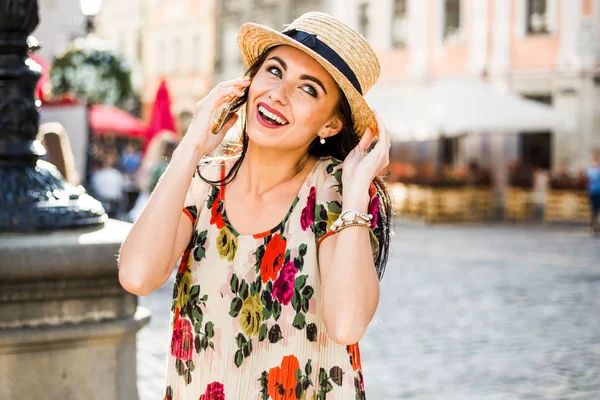 young beautiful girl in summer dress, straw hat. Travels around the European city in the summer. A cheerful, smiling lady is talking on the smartphone and waving her hand. old houses, paving stones