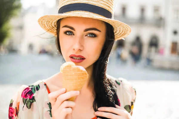 Young beautiful girl in summer dress, straw hat. Travels around the European city in the summer. A cheerful, smiling lady holds ice cream in her hands. Ancient houses, paving stones, vintage style