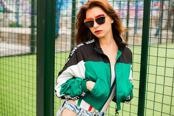 Portrait of a girl clothes in the style of the 90s sports style, jacket, jeans bananas, sunglasses, sports field residential area